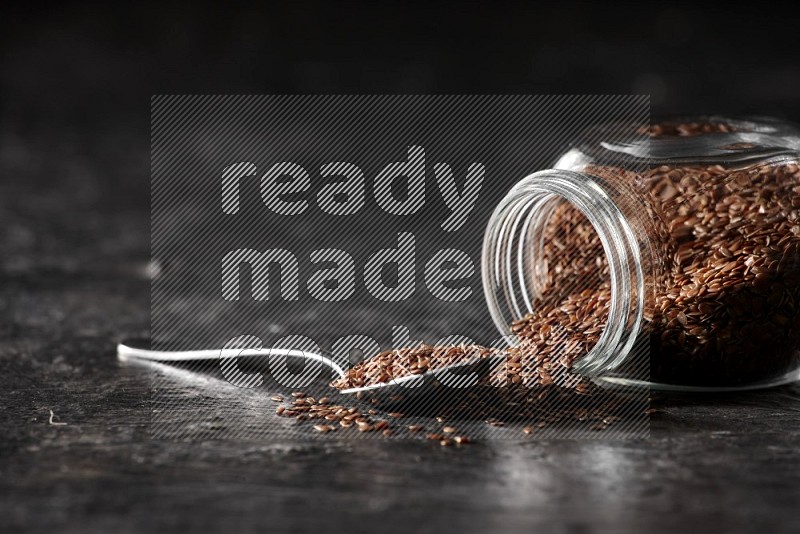 A glass spice jar full of flax flipped and seeds spread out with a metal spoon full of the seeds on a textured black flooring in different angles