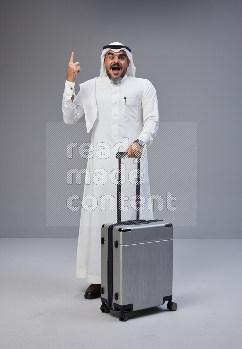 Saudi man wearing Thob and white Shomag standing holding Travel bag on Gray background