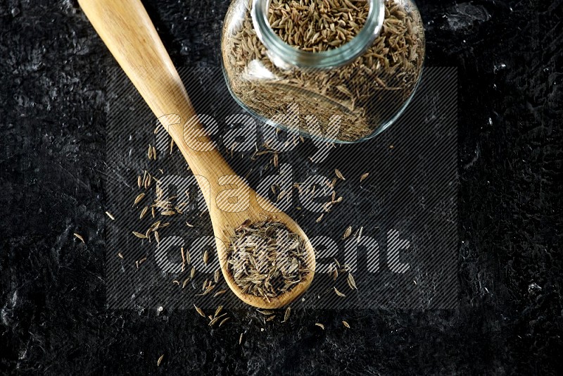 A glass spice jar and a wooden spoon full of cumin seeds on a textured black flooring