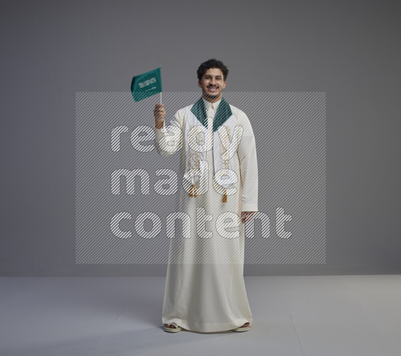 A Saudi man standing wearing thob and saudi flag scarf and holding small saudi flag on gray background