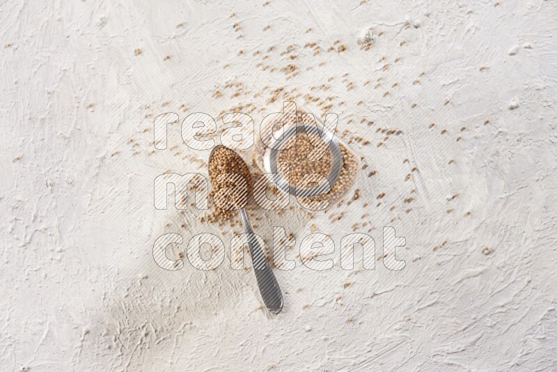 A glass spice jar and a metal spoon full of mustard seeds on a textured white flooring