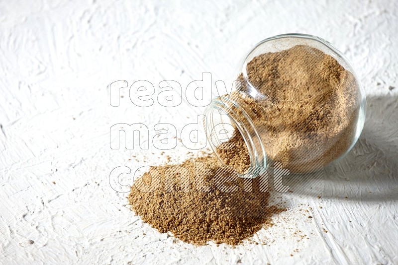 A flipped glass spice jar full of cumin powder and powder spilled out on textured white flooring