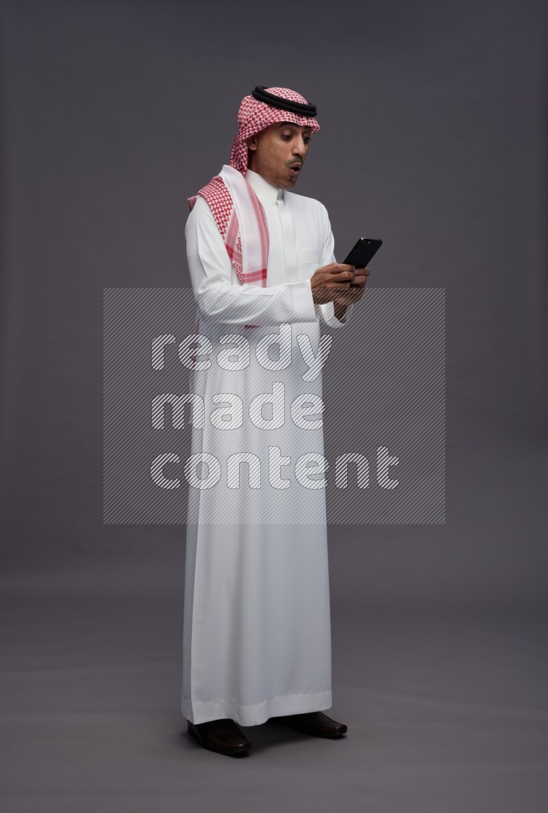 Saudi man wearing thob and shomag standing texting on phone on gray background