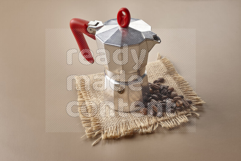 A moka pot with red handle surrounded by roasted coffee beans on beige background
