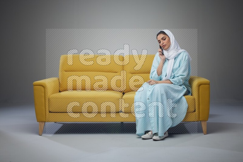 A Saudi woman wearing a light blue Abaya and a white head scarf sitting on a yellow sofa and talking in the phone while making different poses eye level on a grey background
