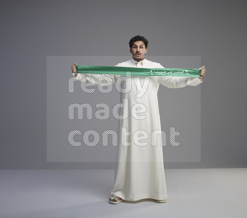 A Saudi man standing wearing thob holding Saudi flag scarf on gray background