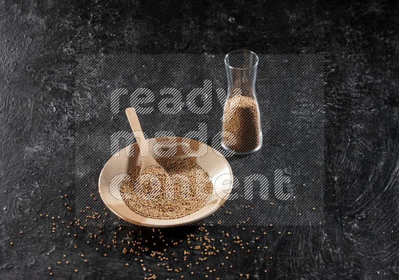 A beige pottery plate full of mustard seeds and a wooden spoon in it with a glass jar filled with the seeds on a textured black flooring