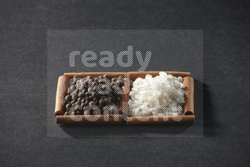 مربعان من أعواد القرفة ممتلئان بالملح الأبيض وحبوب الفلفل الحلو علي خلفية سوداء