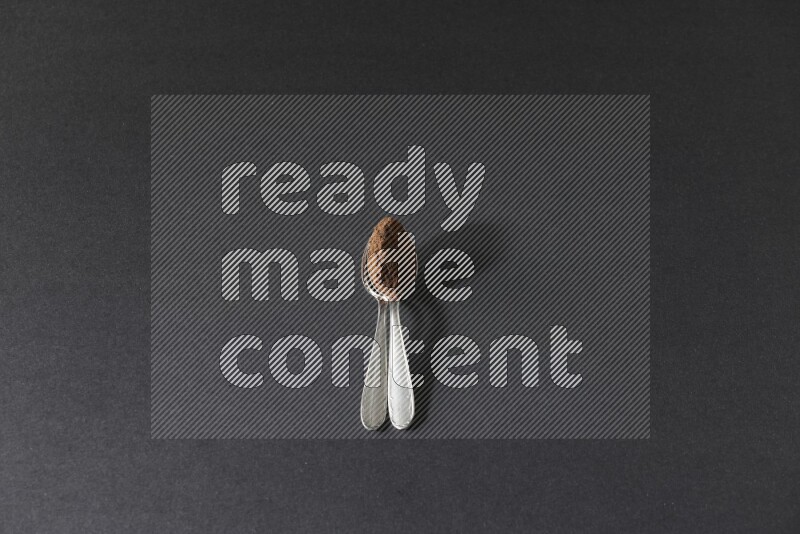 2 metal spoons on top of each other full of cloves powder on a black flooring