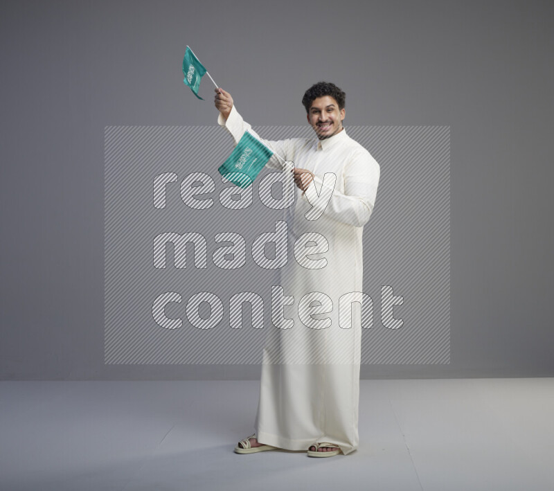A Saudi man standing wearing thob holding small Saudi flag on gray background