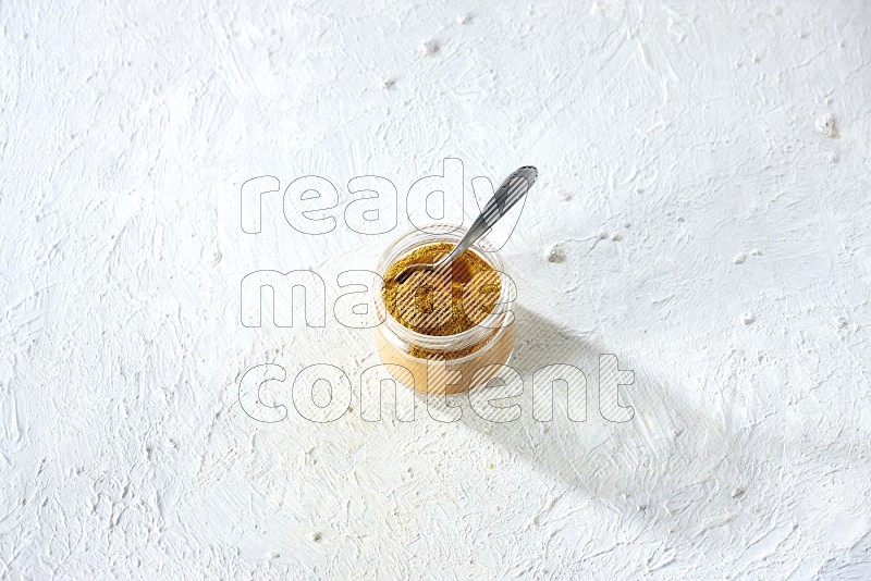 A glass jar and a metal spoon full of turmeric powder on a textured white flooring