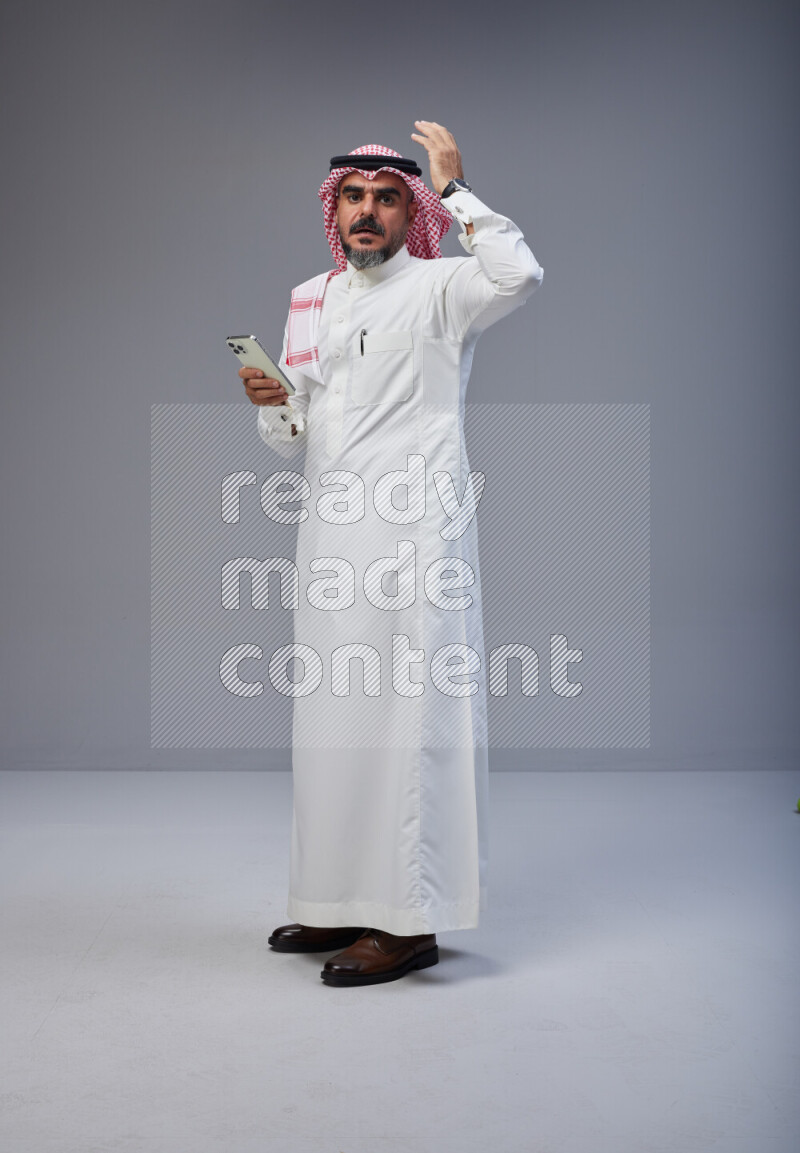 Saudi man Wearing Thob and red Shomag standing texting on phone on Gray background
