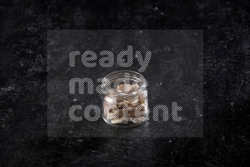 A glass jar full of garlic cloves on a textured black flooring in different angles