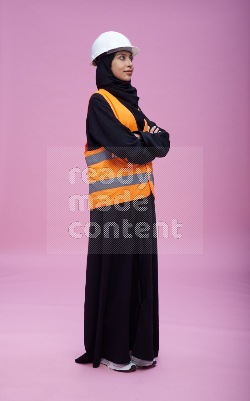 Saudi woman wearing Abaya with engineer vest and helmet standing with crossed arms on pink background
