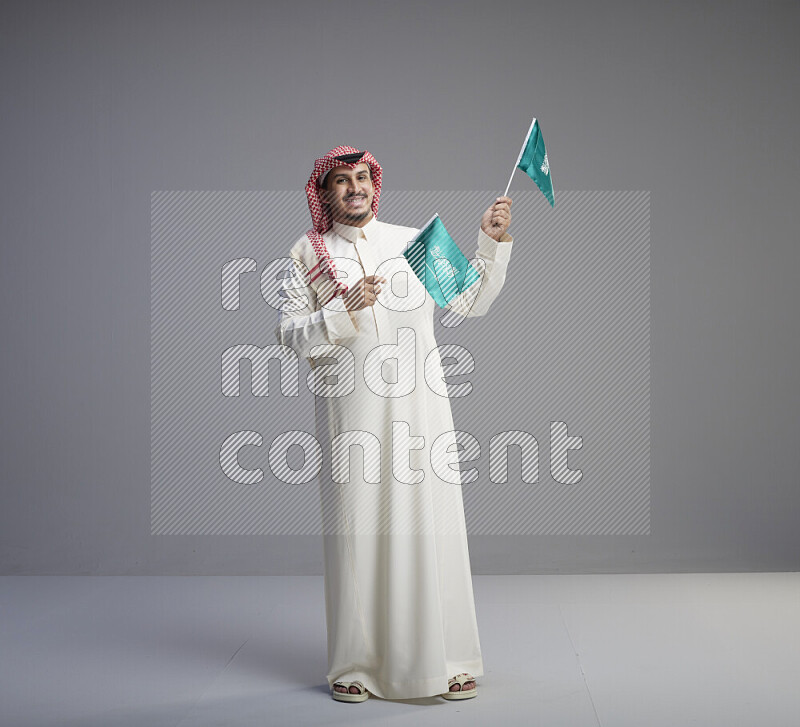 A Saudi man standing wearing thob and red shomag raising small Saudi flag on gray background