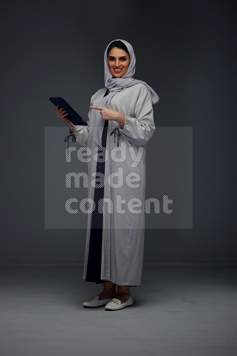 A Saudi woman wearing a light gray Abaya and head scarf standing holding a phone and pointing with the other hand eye level on a grey background