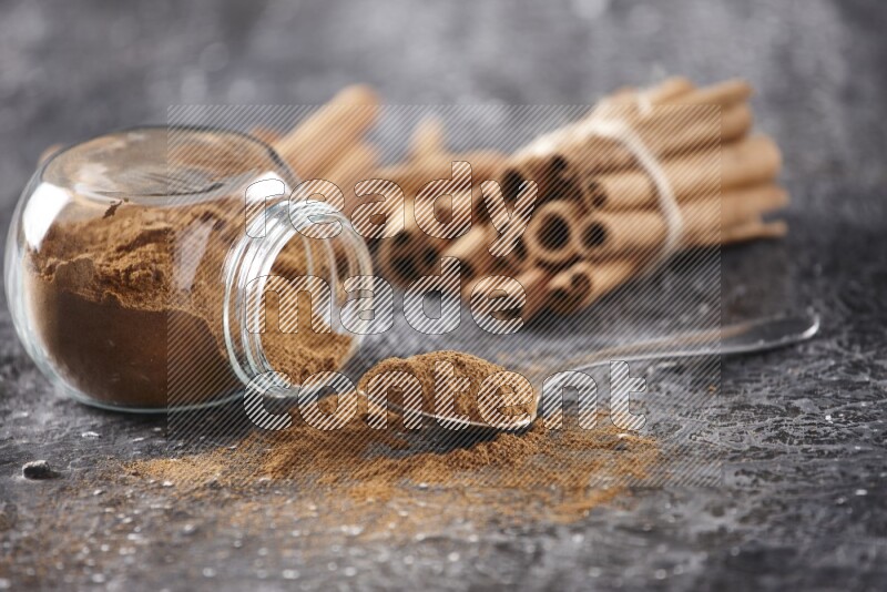 Herbal glass jar full cinnamon powder flipped and a metal spoon full of powder, cinnamon sticks stacked and bounded in the back on textured black background in different angles