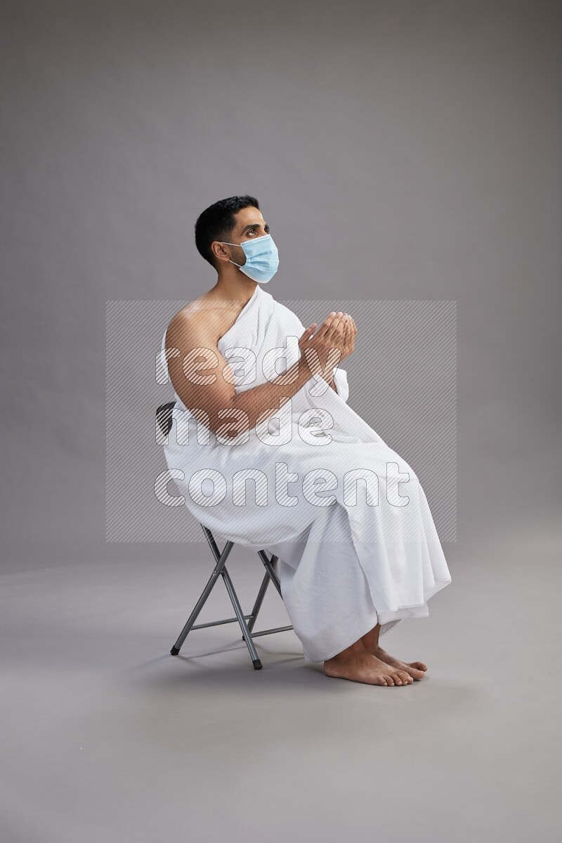 A man wearing Ehram with face mask sitting on chair dua'a on gray background