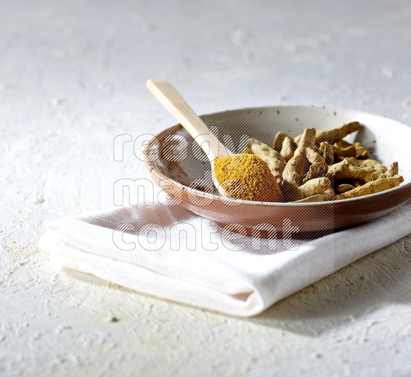 A plate filled with dried turmeric whole fingers and a wooden spoon full of turmeric powder on a textured white flooring