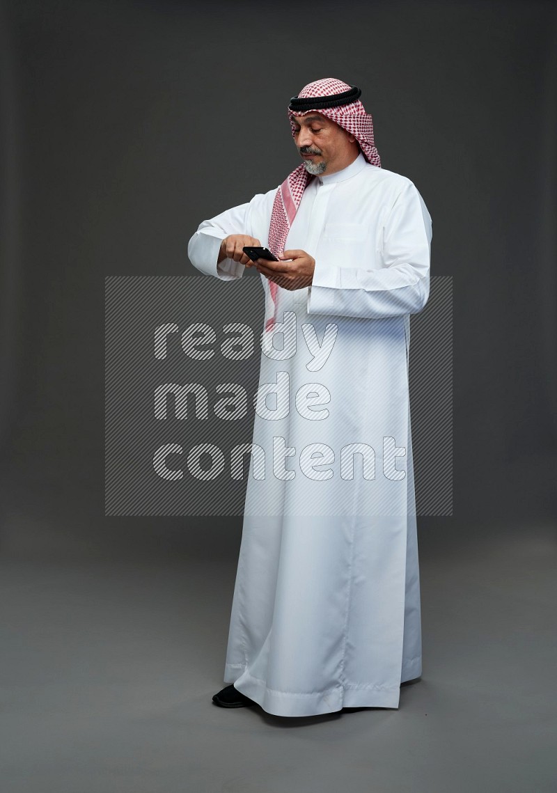 Saudi man with shomag Standing texting on phone on gray background