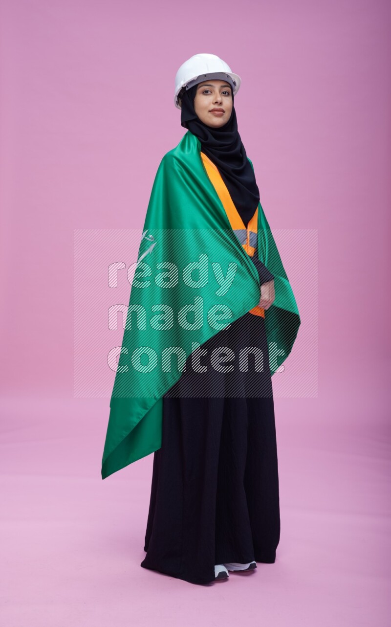 Saudi woman wearing Abaya with engineer vest and helmet standing holding Saudi flag on pink background
