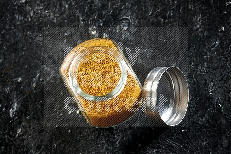 A glass spice jar full of turmeric powder on a textured black flooring