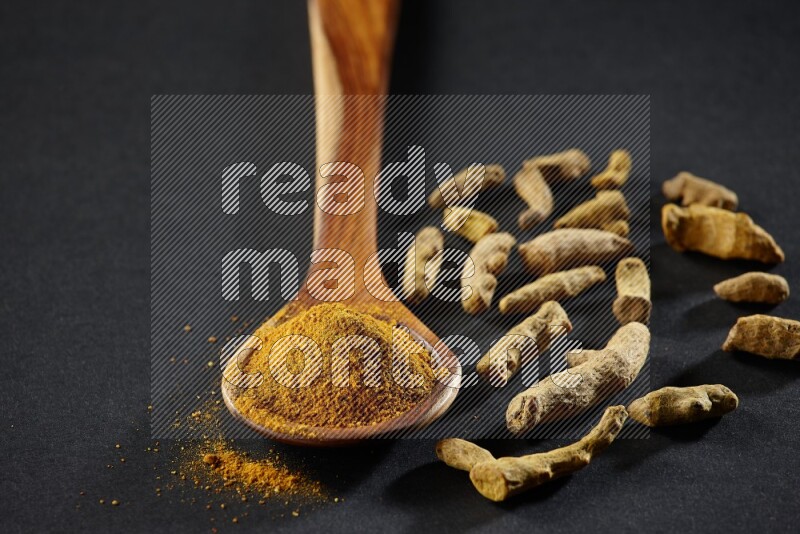 A wooden ladle full of turmeric powder with dried turmeric fingers on black flooring