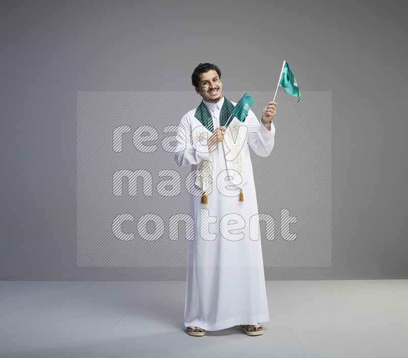 A Saudi man standing wearing thob and saudi flag scarf with face painting holding small Saudi flag on gray background