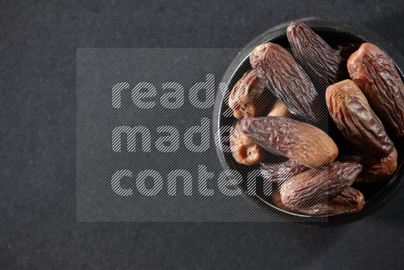 A black pottery bowl full of dried dates on a black background in different angles