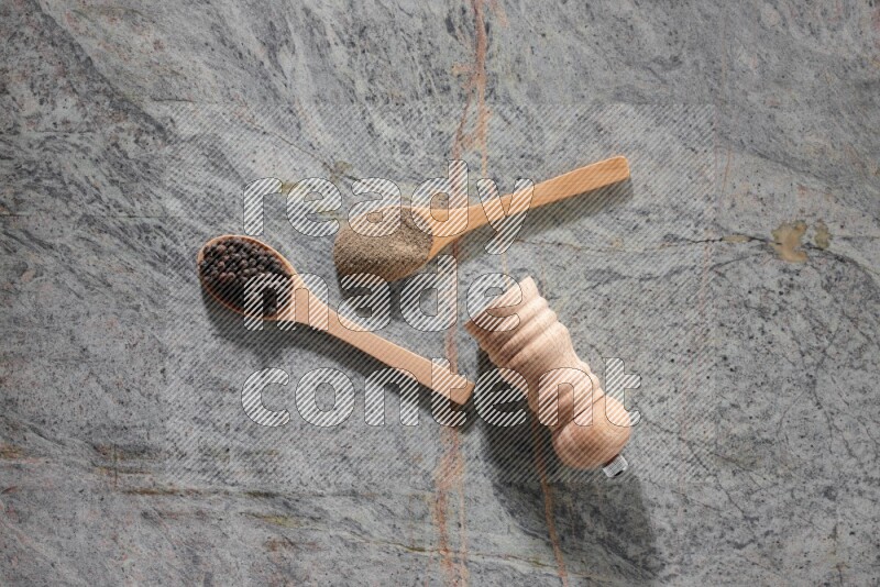 2 wooden spoons, one full of black pepper powder and the other full of black pepper beads and wooden grinder on a marble flooring