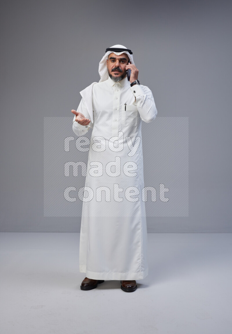Saudi man Wearing Thob and white Shomag standing talking on phone on Gray background