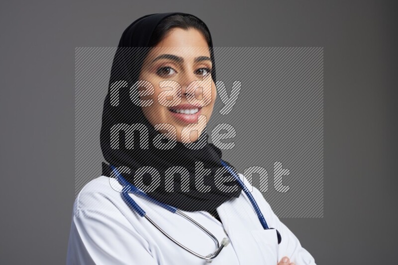 Saudi woman wearing lab coat with stethoscope standing with crossed arms on Gray background