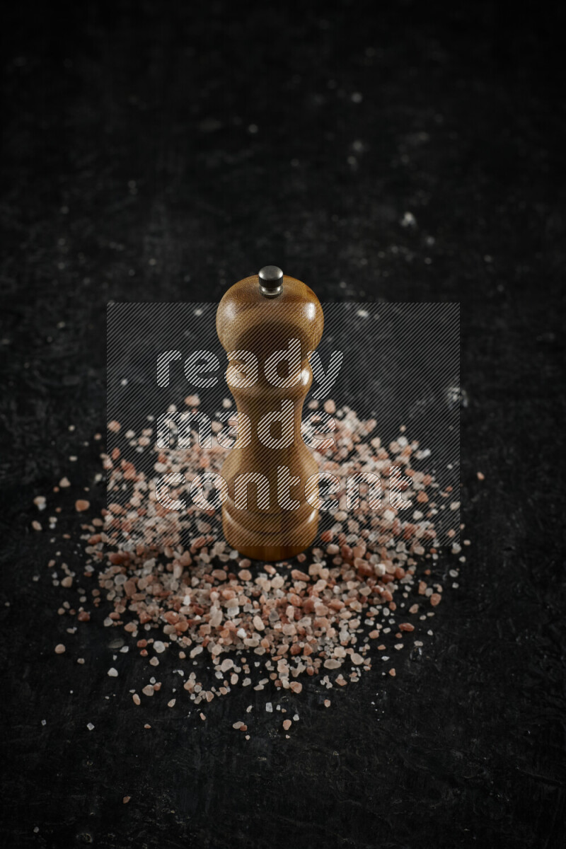 A wooden grinder standing upright and surrounded by coarse pink himalayan salt on black background