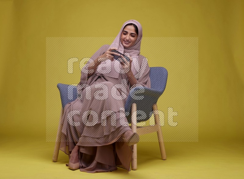 A woman Playing Game Sitting  on a Yellow Background wearing Brown Abaya with Hijab