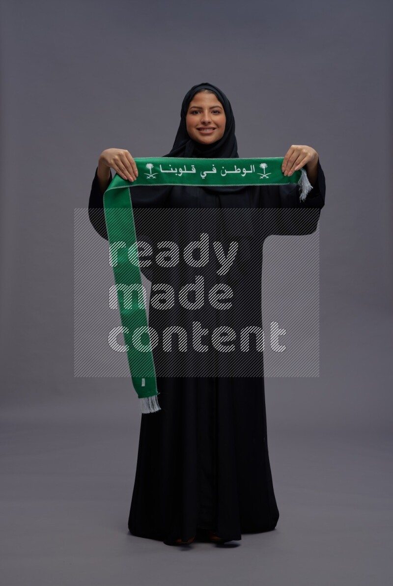 Saudi woman wearing Abaya standing holding Saudi flag on gray background