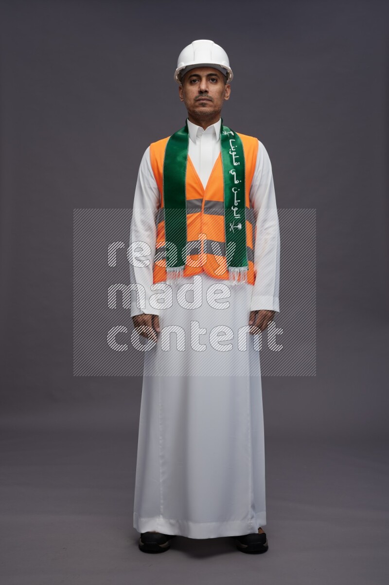 Saudi man wearing thob with engineer vest standing holding Saudi flag on gray background