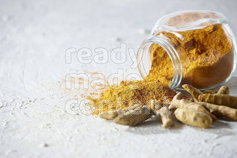 A flipped glass spice jar full of turmeric powder and powder spilled out of it with dried whole fingers on textured white flooring