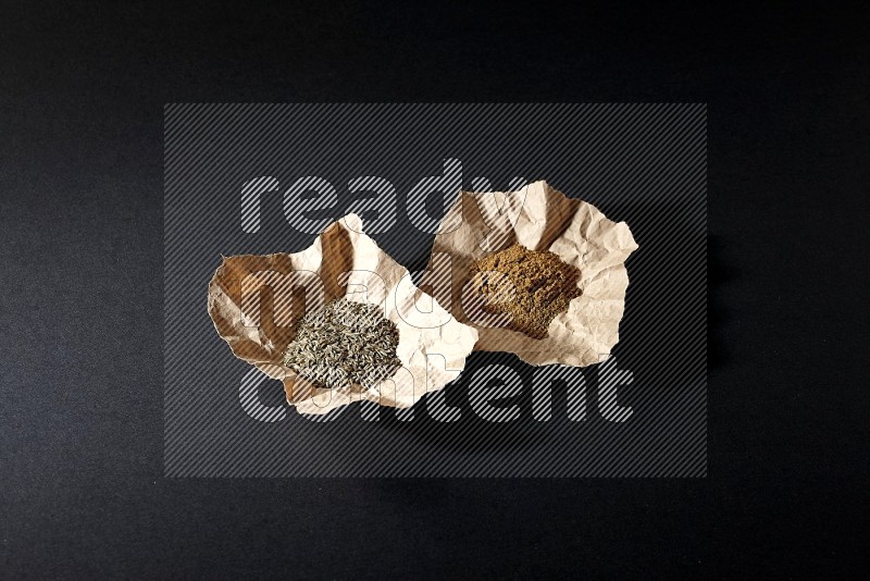 Cumin seeds and powder in 2 crumpled pieces of paper on black flooring