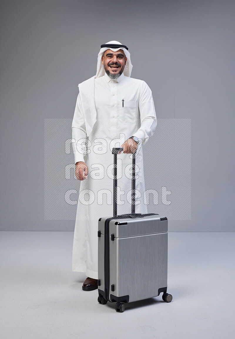 Saudi man wearing Thob and white Shomag standing holding Travel bag on Gray background