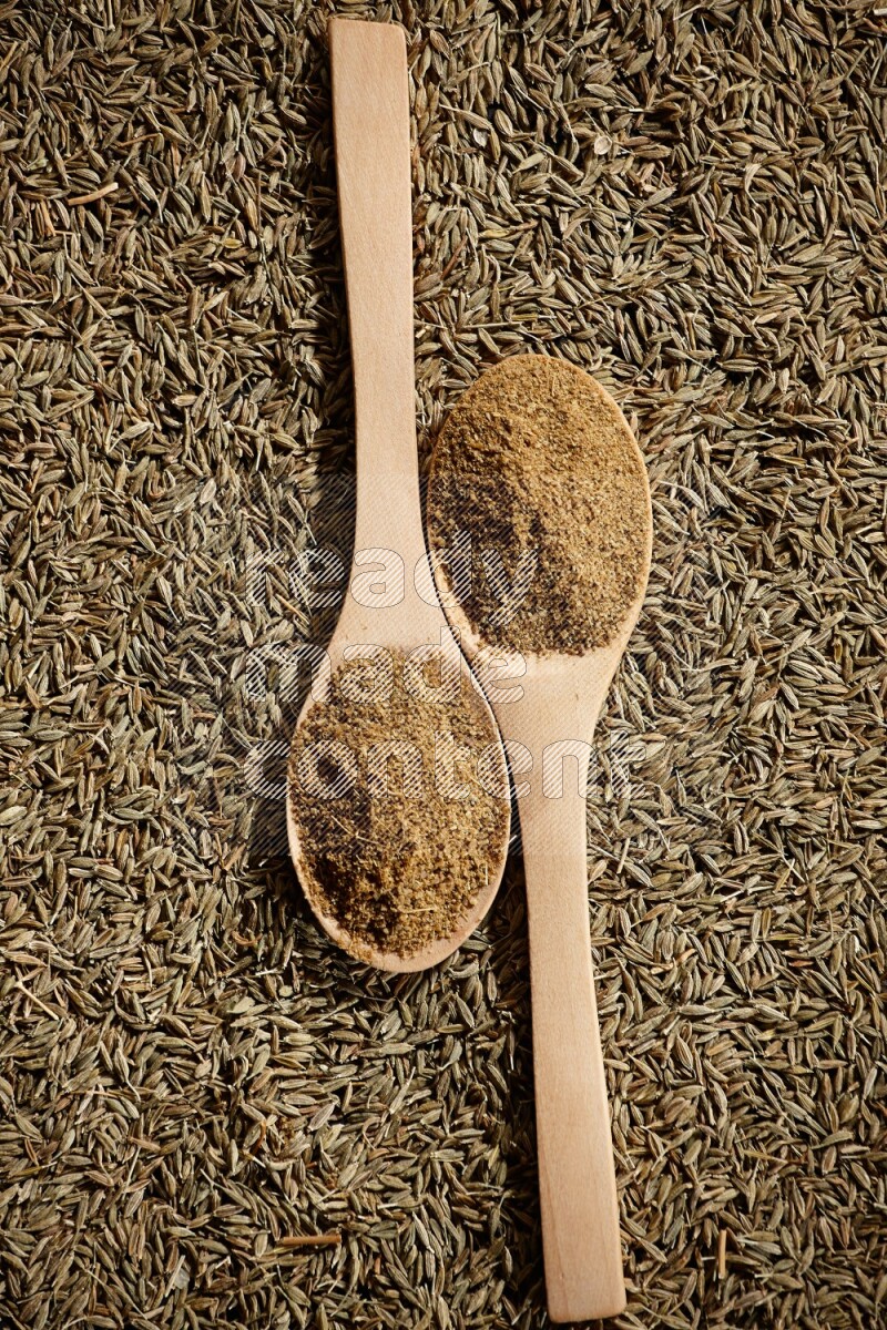 2 wooden spoons full of cumin powder on a cumin seeds background