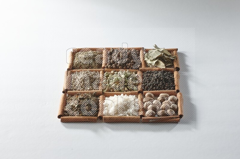 9 squares of cinnamon sticks full of cardamom in the middle surrounded by nutmeg, salt, dried basil, white pepper, cumin, allspice, bay laurel leaves and black pepper on white flooring