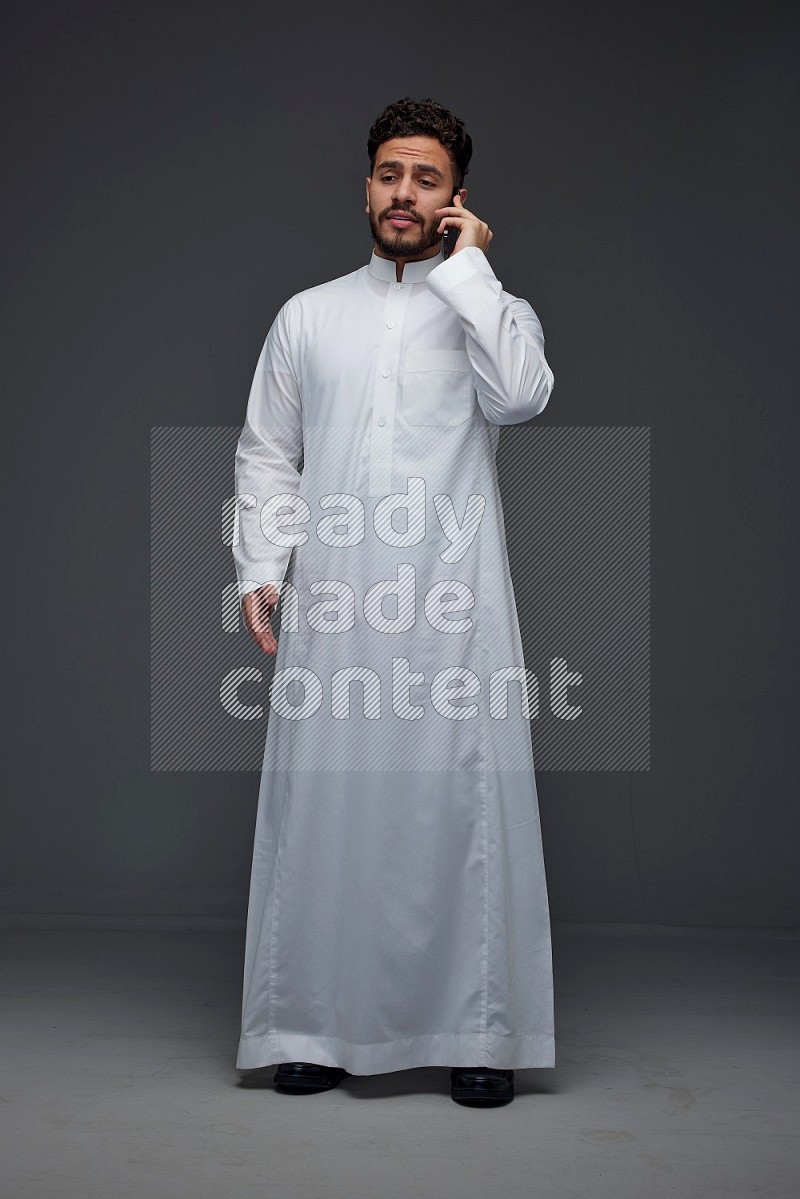 A Saudi man wearing Thobe and talking in the phone while standing and making different hand gestures eye level on a gray background