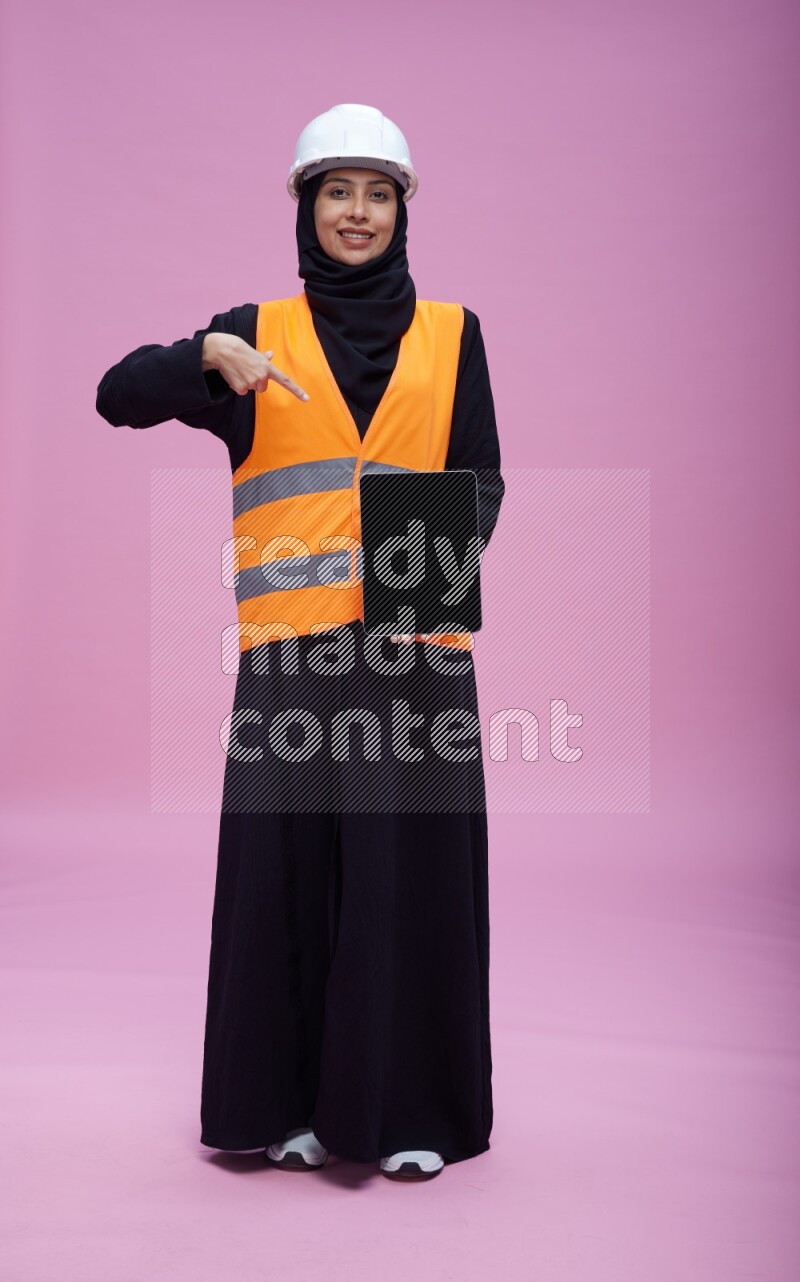 Saudi woman wearing Abaya with engineer vest and helmet standing showing tablet to camera on pink background