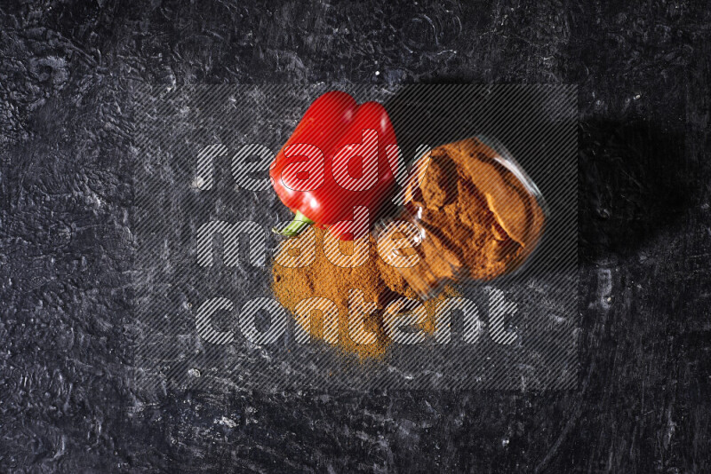 A glass jar full of ground paprika powder flipped with some spilling powder on black background