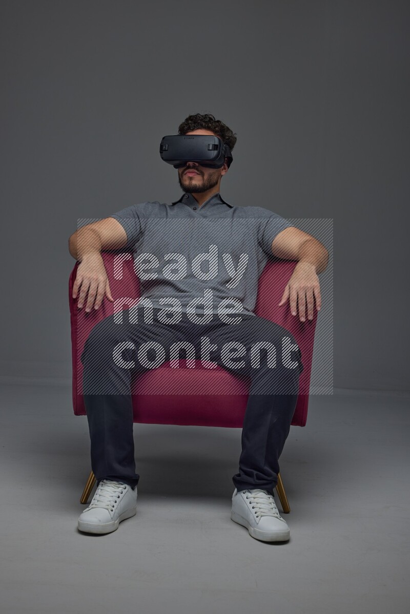 A man wearing casual and wearing VR while sitting on a burgundy chair eye level on a gray background