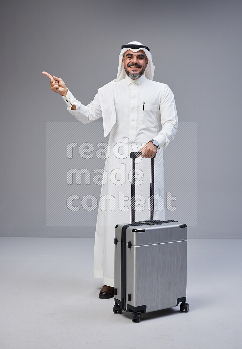 Saudi man wearing Thob and white Shomag standing holding Travel bag on Gray background