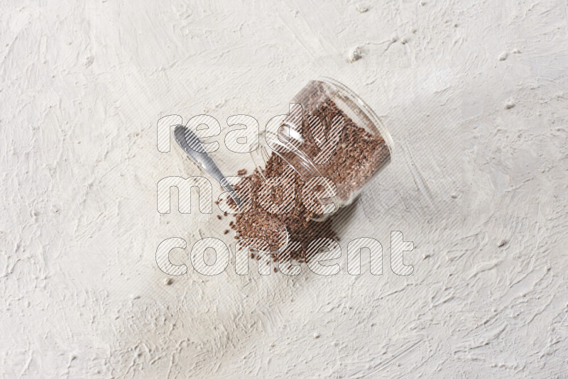 A glass spice jar full of flax seeds flipped with a metal spoon full of the seeds on a textured white flooring