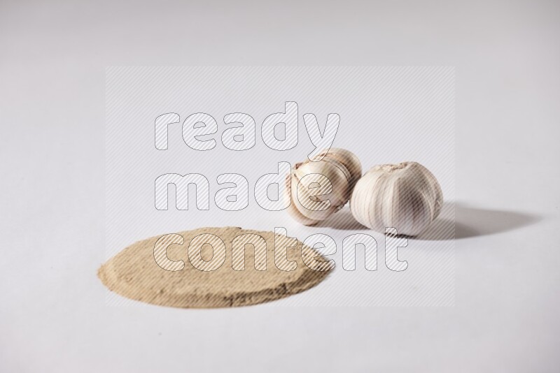 Garlic powder in a circle shape and beside it 2 garlic bulbs on a white flooring