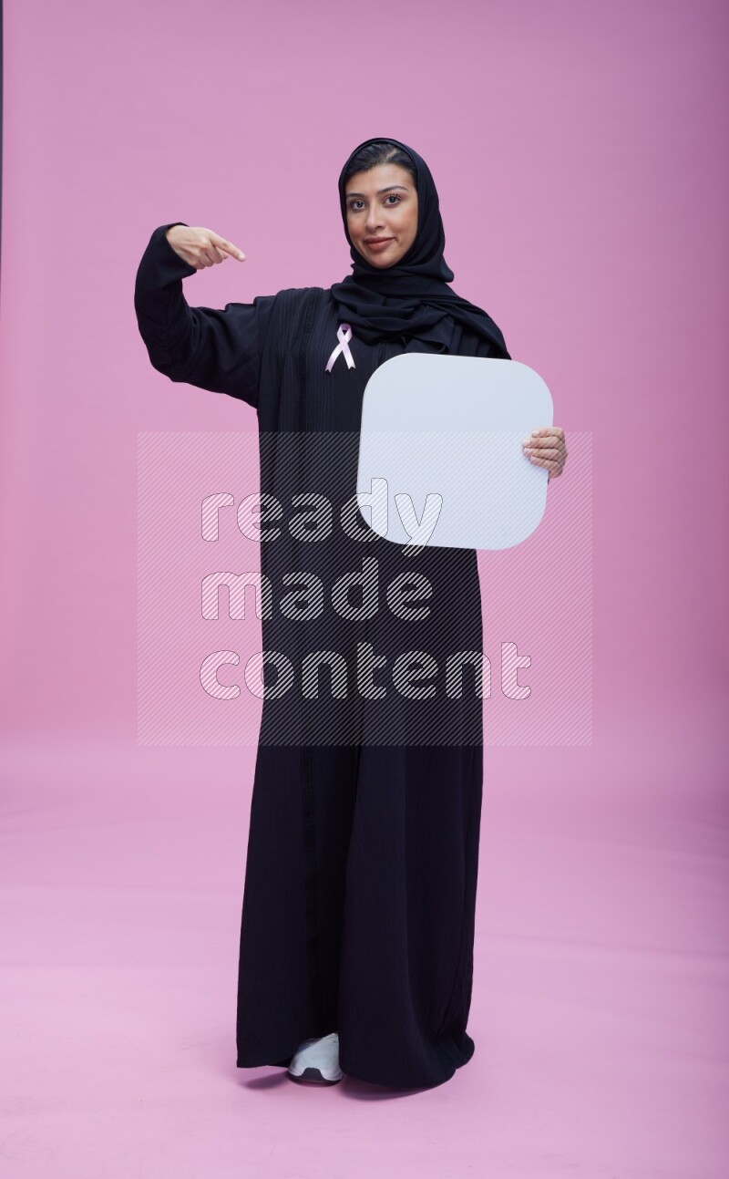 Saudi woman wearing pink ribbon on Abaya standing holding social media signs on pink background