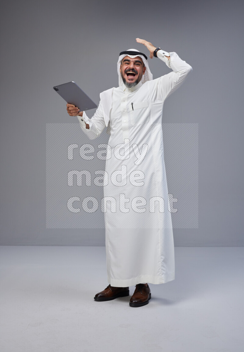 Saudi man Wearing Thob and white Shomag standing working on tablet on Gray background
